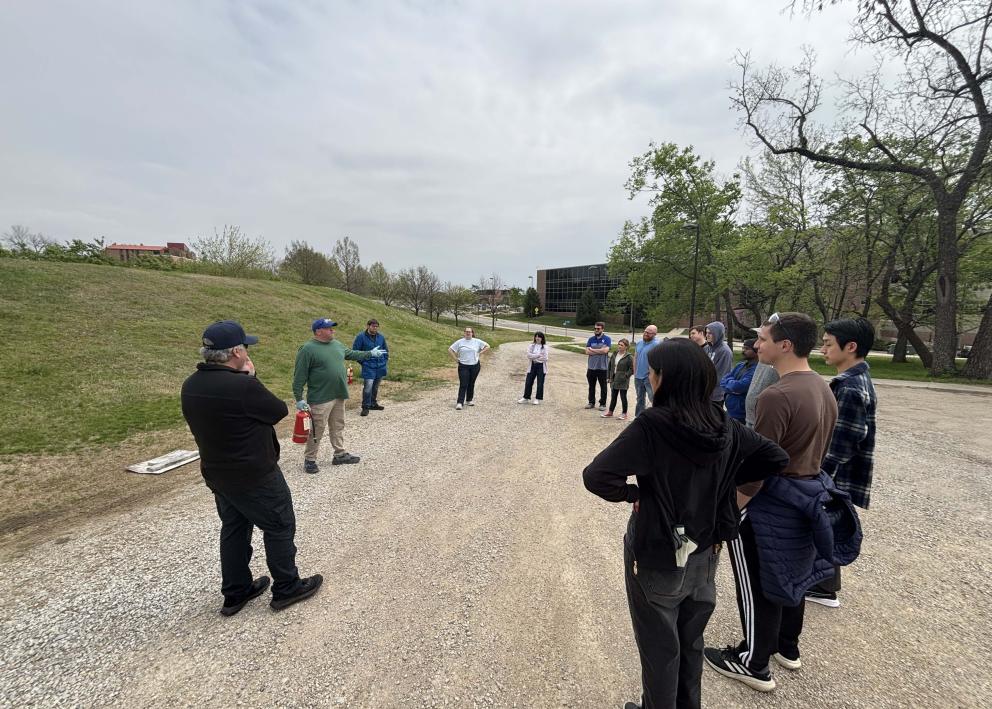 A group of people gathered on a gravel road with one person speaking holding a fire extinguisher