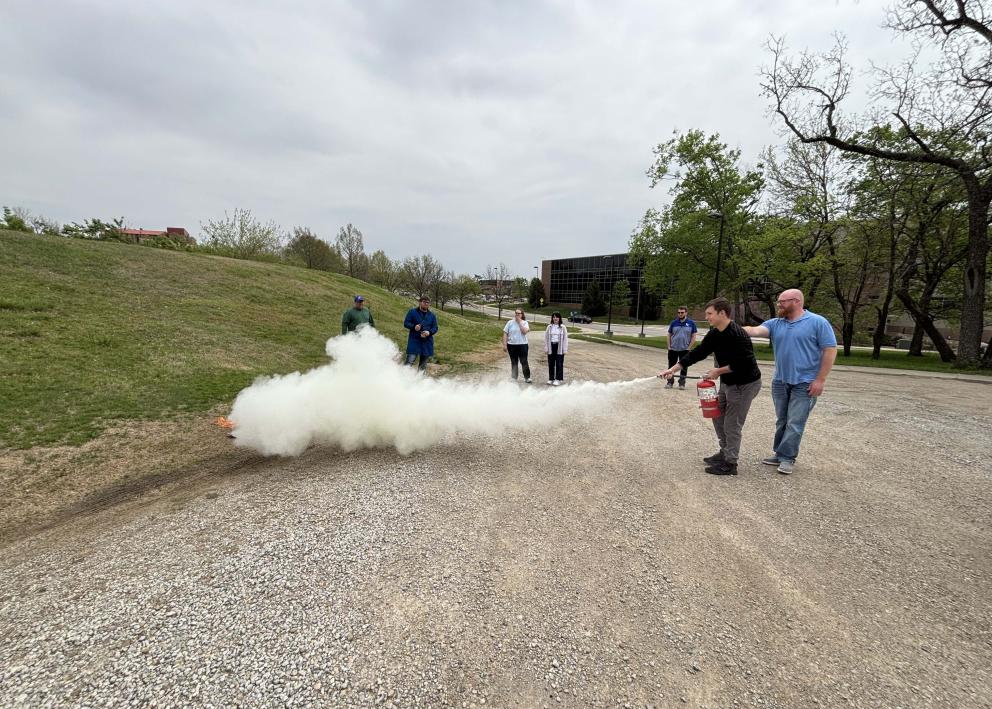 A group of people on a gravel road, one is using a fire extinguisher to put out a small fire on the side of the road