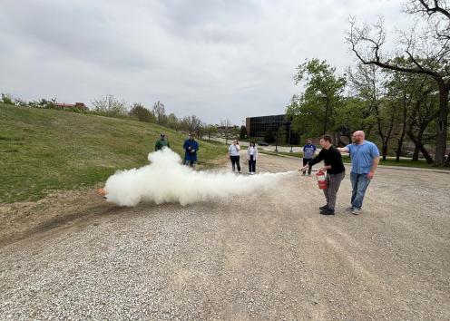 A group of people on a gravel road, one is using a fire extinguisher to put out a small fire on the side of the road